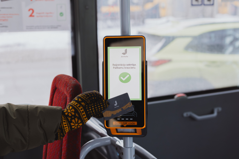 Autobusa validātors ar oranžu rāmi, uz kura cilvēks ar melniem-dzelteniem rakstainiem cimdiem pieskaras ar karti — ekrānā zaļš ķeksītis