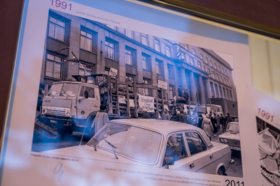 Melnbalta fotogrāfija no 1991. gada: pie valdības ēkas redzamas barikādes ar kravas automašīnām, kas piekrautas ar baļķiem, apkārt pulcējušies cilvēki; attēls ir daļa no izstādes stenda par Barikāžu laiku