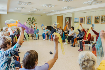 Seniori sasēdušies aplī vingro un tur lakatiņus rokās