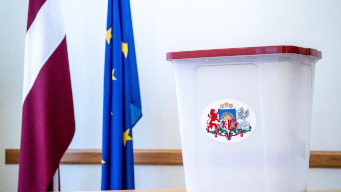 Vēlēšanu urna ar Latvijas ģerboni uz tās, novietota uz galda; fonā redzams Latvijas un Eiropas Savienības karogs