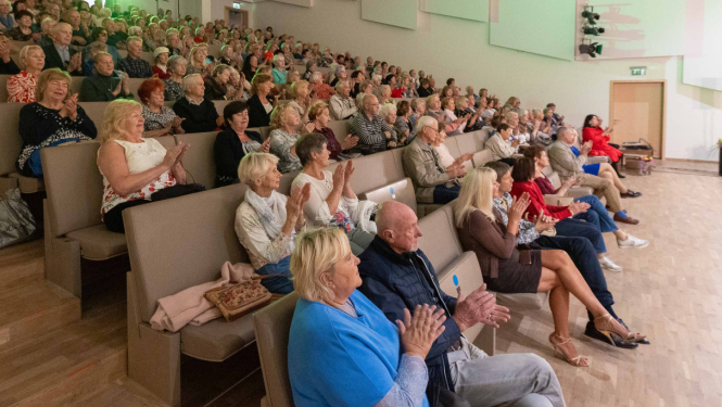 Liela auditorija ar sēdošiem skatītājiem, galvenokārt senioriem, aplausē koncerta laikā