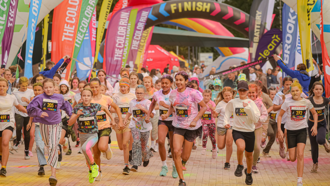 Grupa bērnu un pusaudžu entuziasma pilni startē krāsainā skriešanas pasākumā 'The Color Run', uzsākot skrējienu zem finiša arkas, apkārt ir daudzkrāsaini karogi un skatītāji