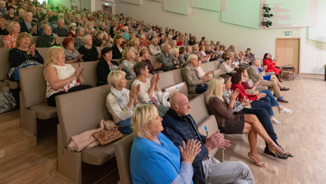 Liela auditorija ar sēdošiem skatītājiem, galvenokārt senioriem, aplausē koncerta laikā