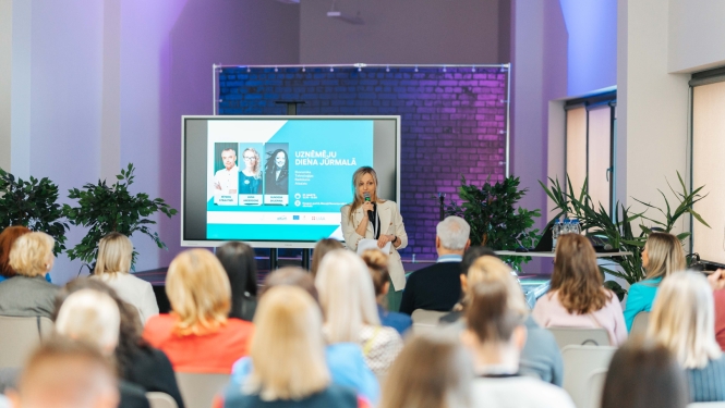 Konference ar nosaukumu 'Uzņēmēju diena Jūrmalā', kurā sieviete ar mikrofonu runā auditorijas priekšā