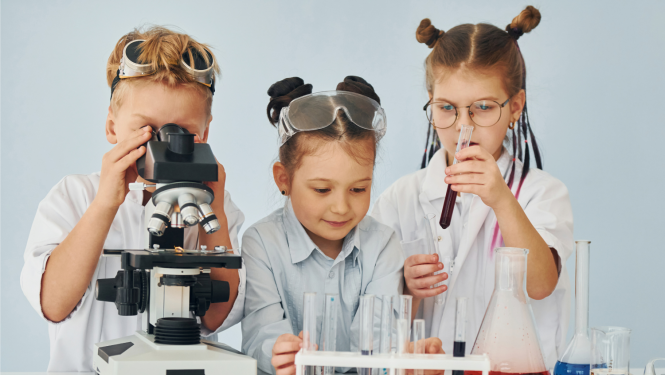 Trīs bērni laboratorijā veic zinātniskus eksperimentus — viens aplūko paraugu mikroskopā, bet pārējie strādā ar mēģenēm un laboratorijas traukiem.