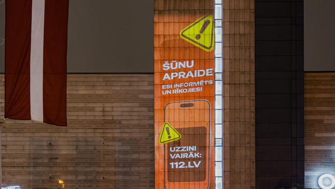 Ēkas fasāde ar izgaismotu vertikālu reklāmu oranžā krāsā, kurā redzams brīdinājuma simbols un teksts latviešu valodā: ‘Šūnu apraide. Esi informēts un rīkojies! Uzzini vairāk: 112.lv