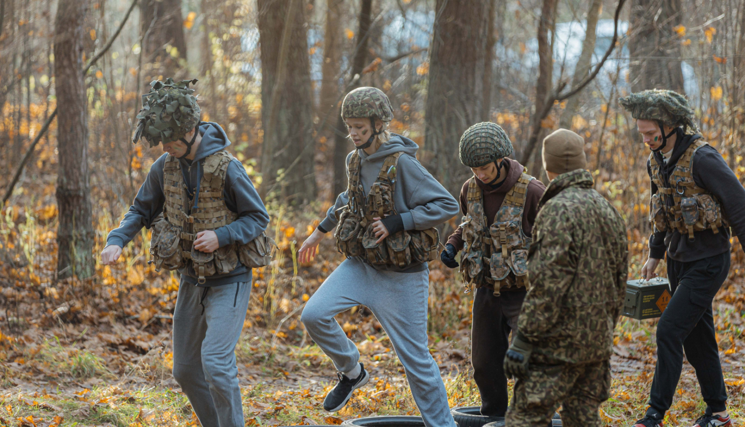 Jaunieši militārā tipa tērpos un ķiverēs piedalās orientēšanās un veiklības uzdevumā mežā, soļojot cauri riepu šķēršļiem instruktora uzraudzībā