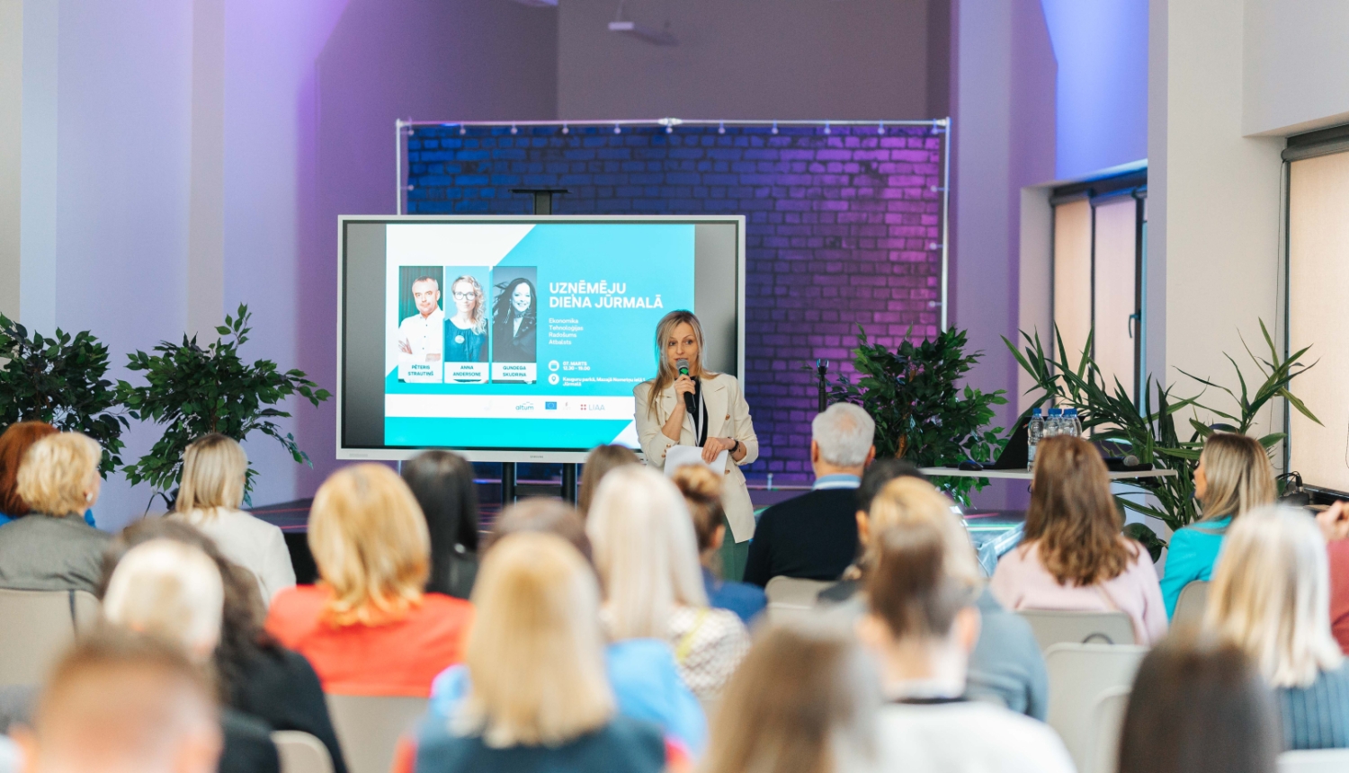 Konference ar nosaukumu 'Uzņēmēju diena Jūrmalā', kurā sieviete ar mikrofonu runā auditorijas priekšā
