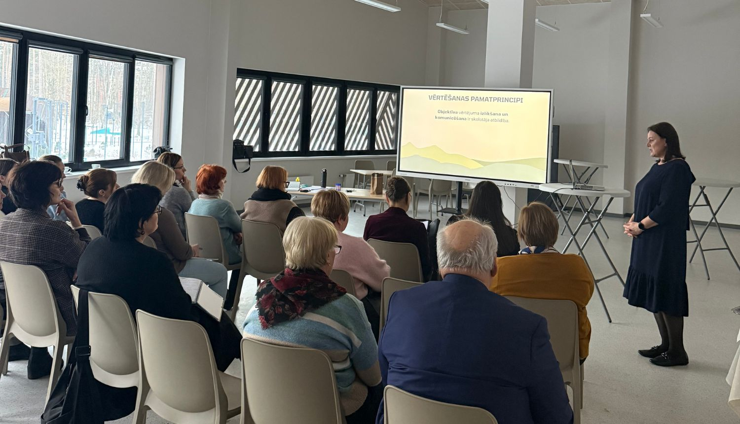 Pedagogu profesionālās pilnveides nodarbība – skolotāji sēž auditorijā un klausās lekciju, ekrānā redzama prezentācija par vērtēšanas pamatprincipiem