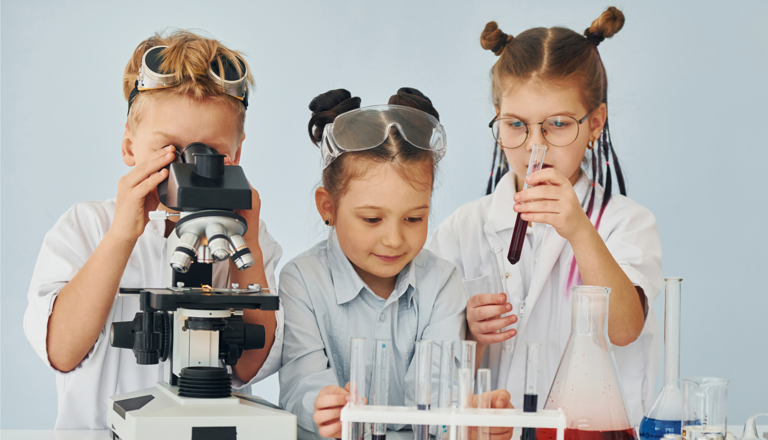 Trīs bērni laboratorijā veic zinātniskus eksperimentus — viens aplūko paraugu mikroskopā, bet pārējie strādā ar mēģenēm un laboratorijas traukiem.