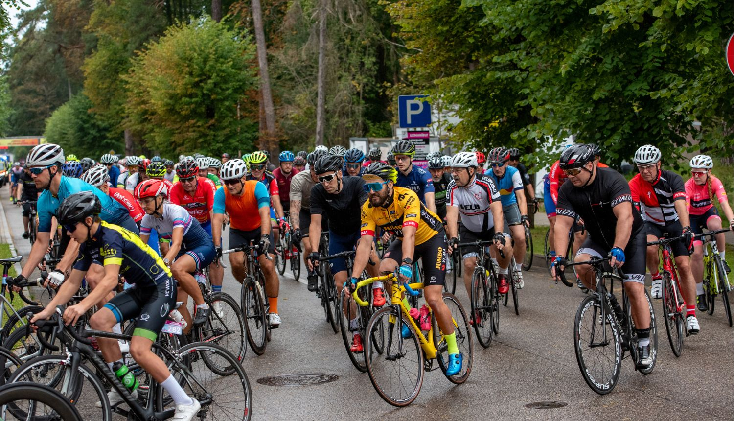 Liela velobraucēju grupa sacensību laikā brauc pa ielu, visi aprīkoti ar ķiverēm un sporta apģērbu