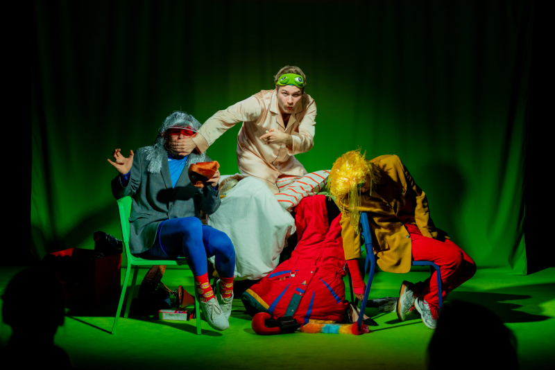 trīs aktieri teātra izrādes ainā ar spilgtiem, komiski pārspīlētiem tērpiem un parūkām, sēžot krāsainos krēslos uz zaļa fona