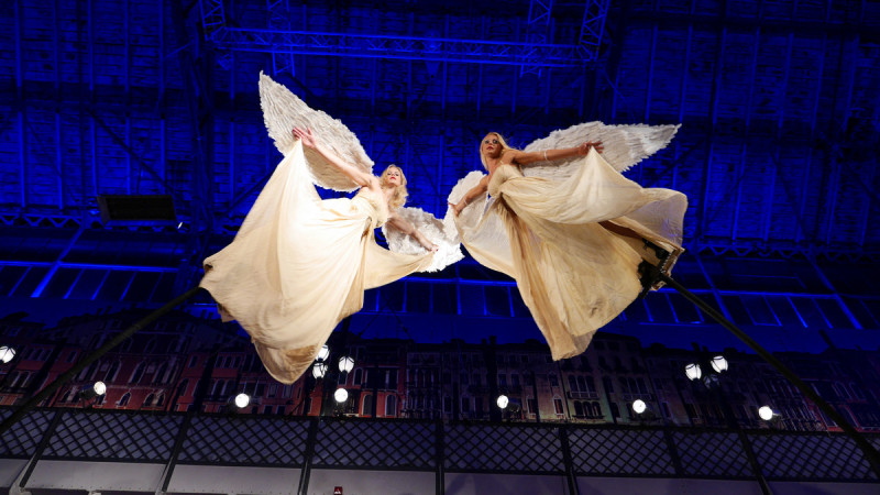 divas dejotājas gaisā, tērpušās garās, plūstošās krēmkrāsas kleitās ar lieliem baltiem spārniem, kas atgādina eņģeļus