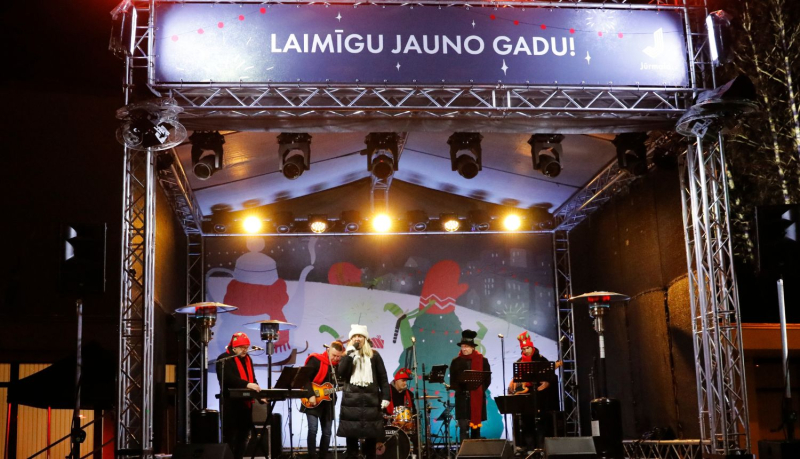Jaunā gada sagaidīšana Kauguros, kurā redzama skatuve ar dziedātājiem un uzrakstu Laimīgu Jauno gadu