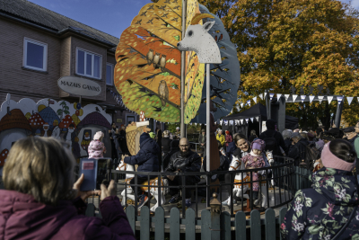 Cilvēki svētku pasākumā brīvā dabā, kur bērni brauc ar mazu karuseli, kas rotāts ar krāsainām koka figūrām — zirgiem, dzīvniekiem un rudens lapu motīviem