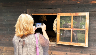 Sieviete ar gaišiem matiem, mugurā pelēks apmetnis un plecā rozā soma, fotografē ar viedtālruni caur nelielu atvērtu logu senlaicīgā koka ēkā