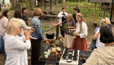 Āra pasākuma laikā grupa cilvēku pulcējas ap ugunskuru, virs kura vārās katls