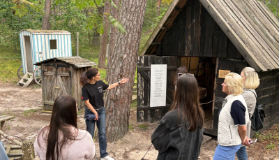 Ekskursijas laikā gide melnā kreklā stāsta grupai cilvēku par nelielu koka ēku ar stāvu jumtu