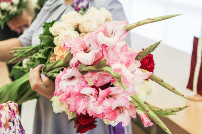 Cilvēks tur rokās krāšņu ziedu pušķi ar rozā gladiolām un krēmkrāsas rozēm – svinīgs un sirsnīgs mirklis apbalvošanas vai sveikšanas pasākumā