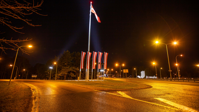 Nakts skats Jūrmalā: apgaismots satiksmes aplis ar paceltu Latvijas valsts karogu un vairākām mazākām karogmastiem svētku noformējumā pie Jūrmalas pilsētas robežas zīmes