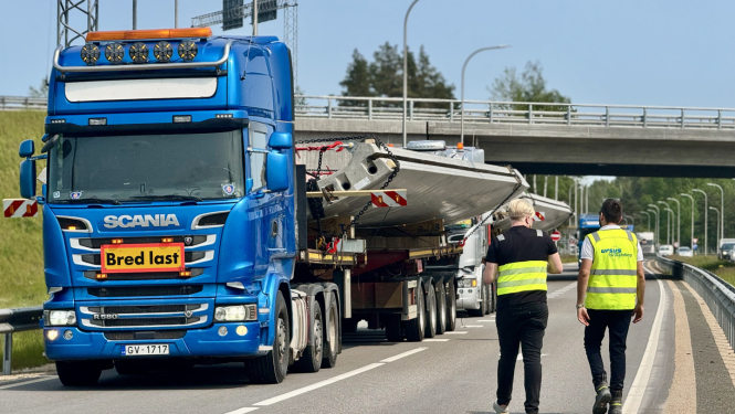 Attēlā redzama zila Scania kravas automašīna ar piekabi, kas pārvadā lielu, platu kravu – iespējams, metāla vai betona konstrukciju. Kravas priekšā ir dzeltena zīme ar sarkanu apmali un uzrakstu "Bred last", kas zviedru valodā nozīmē "Plata krava". Divi vīrieši ar atstarojošām vestēm, viens no kuriem pārstāv uzņēmumu "Ursus Transport", iet gar kravas automašīnu pa ceļa nomali. Fonā redzama automaģistrāle ar vairākām joslām, tilts un citas automašīnas. Attēls ataino kravas pārvadājumu procesa pārraudzību vai