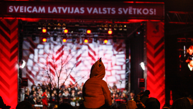 Svētku koncerts brīvdabas skatuvē ar sarkanbaltiem gaismu efektiem un uzrakstu “Sveicam Latvijas valsts svētkos!”, skatītāju pūlī redzams bērns uz pieaugušā pleciem