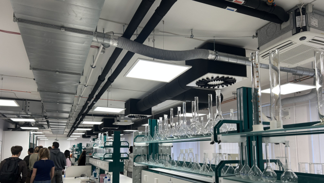 Skolēni ekskursijā laboratorijā, kur garos darba galdos izvietoti stikla mēģenes, kolbas un cita laboratorijas stikla trauki