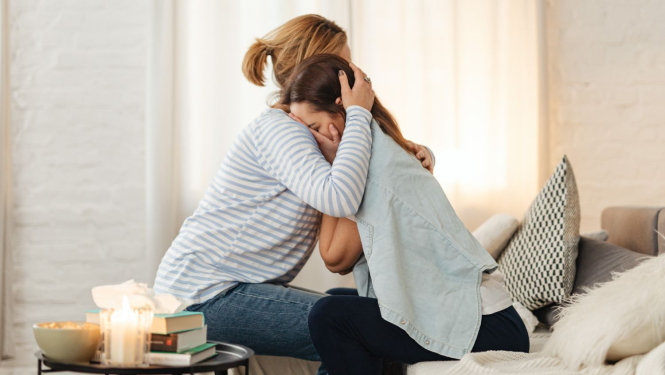 sieviete apskauj otru sievieti, kura ir noskumusi