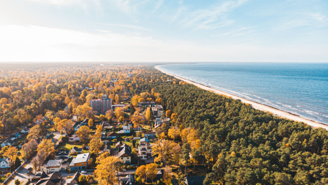 Putna lidojuma skats uz Jūrmalas piekrasti rudenī — redzami koki ar dzeltenīgām lapām, dzīvojamās mājas un daudzstāvu ēka, kā arī pludmale un Baltijas jūra.