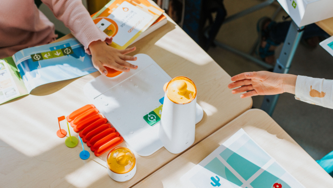 izglītojošs robotikas komplekts uz galda, kas ietver krāsainus elementus, programmēšanas kartītes un grāmatas. Redzamas bērnu rokas, kas mijiedarbojas ar materiāliem saules gaismā apgaismotā klasē