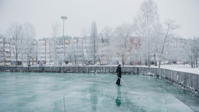 Cilvēks tumšā ziemas apģērbā stāv uz aizsalušas āra slidotavas un ar šļūteni lej ūdeni uz ledus