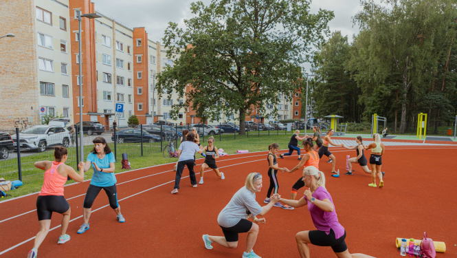 Āra vingrošanas nodarbība uz sarkanā skrejceļa, kurā vairāki sieviešu pāri veic vingrinājumus pa pāriem