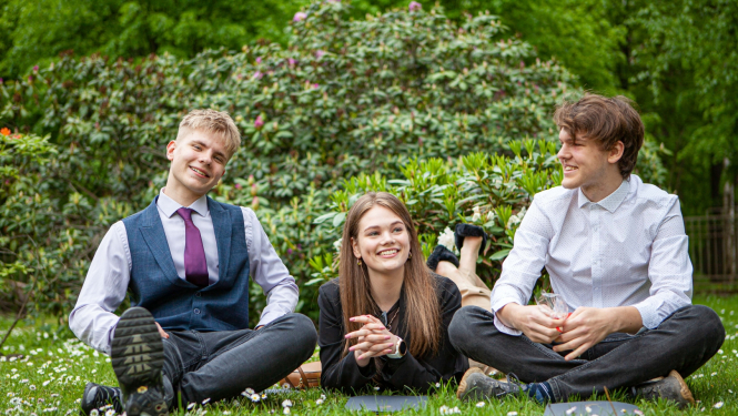 Divi zēni un viena meitene no Pumpuru vidusskolas sēž zaļā zālē pēc liecību saņemšanas