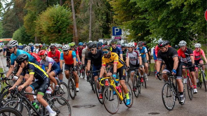 Liela velobraucēju grupa sacensību laikā brauc pa ielu, visi aprīkoti ar ķiverēm un sporta apģērbu