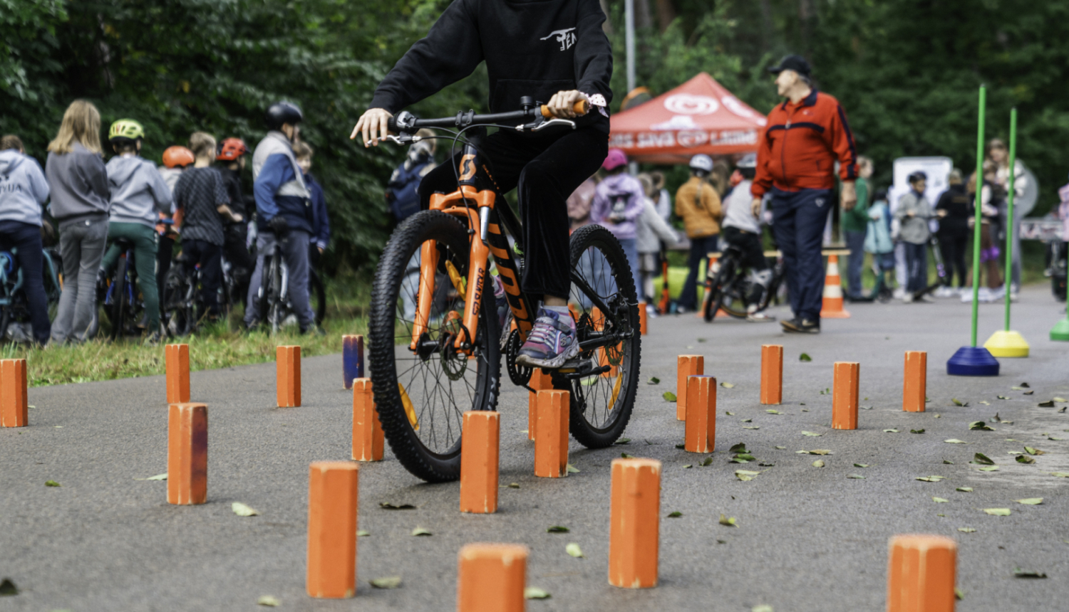 Bērns brauc ar oranžu kalnu velosipēdu cauri šķēršļu trasei, kur izvietoti koši oranži stabiņi uz asfalta seguma