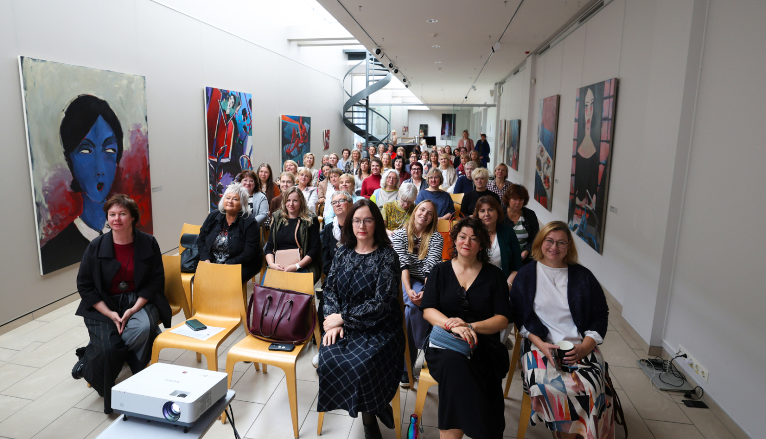 Liela cilvēku grupa sēž krēslos modernā, gaišā izstāžu zālē ar augstiem griestiem