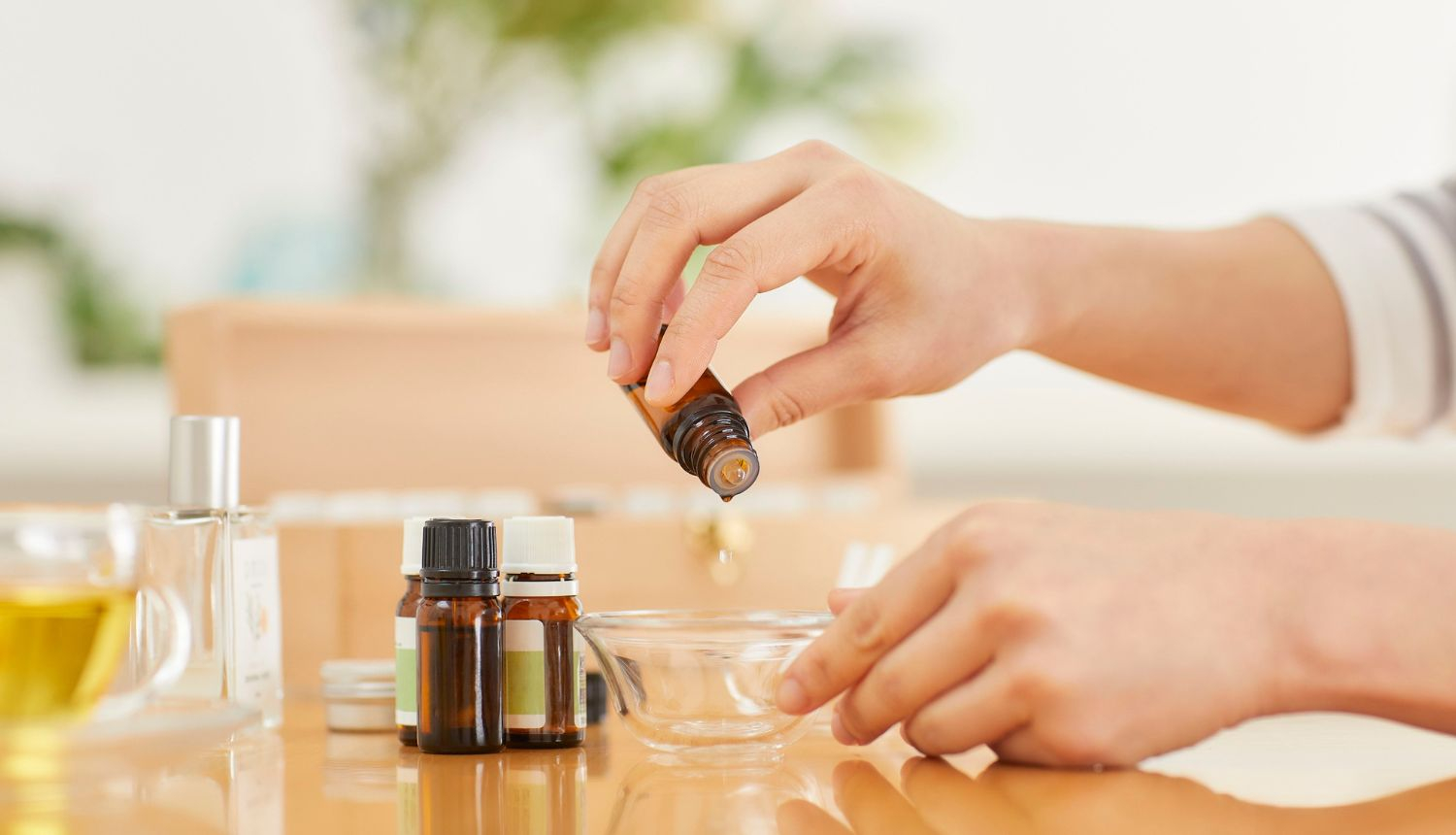 Tuvplāns ar cilvēka rokām, kas iepilina ēterisko eļļu no brūnas stikla pudelītes caurspīdīgā stikla trauciņā. Uz galda redzamas arī citas eļļas pudelītes, smaržūdens flakons un krūze ar zāļu tēju