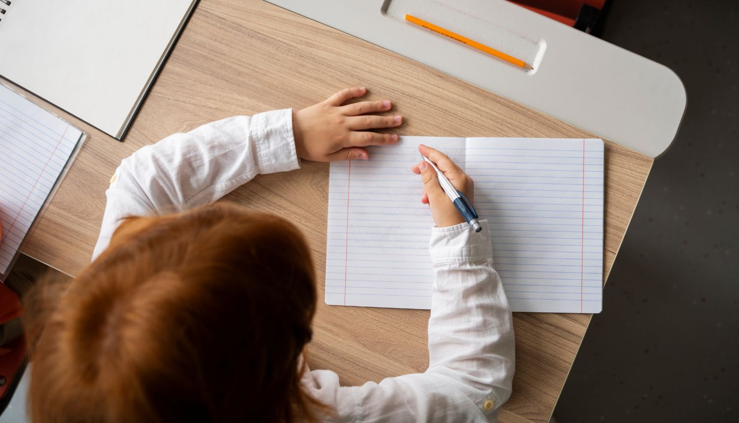 Bērns sēž pie galda un raksta burtnīcā ar pildspalvu
