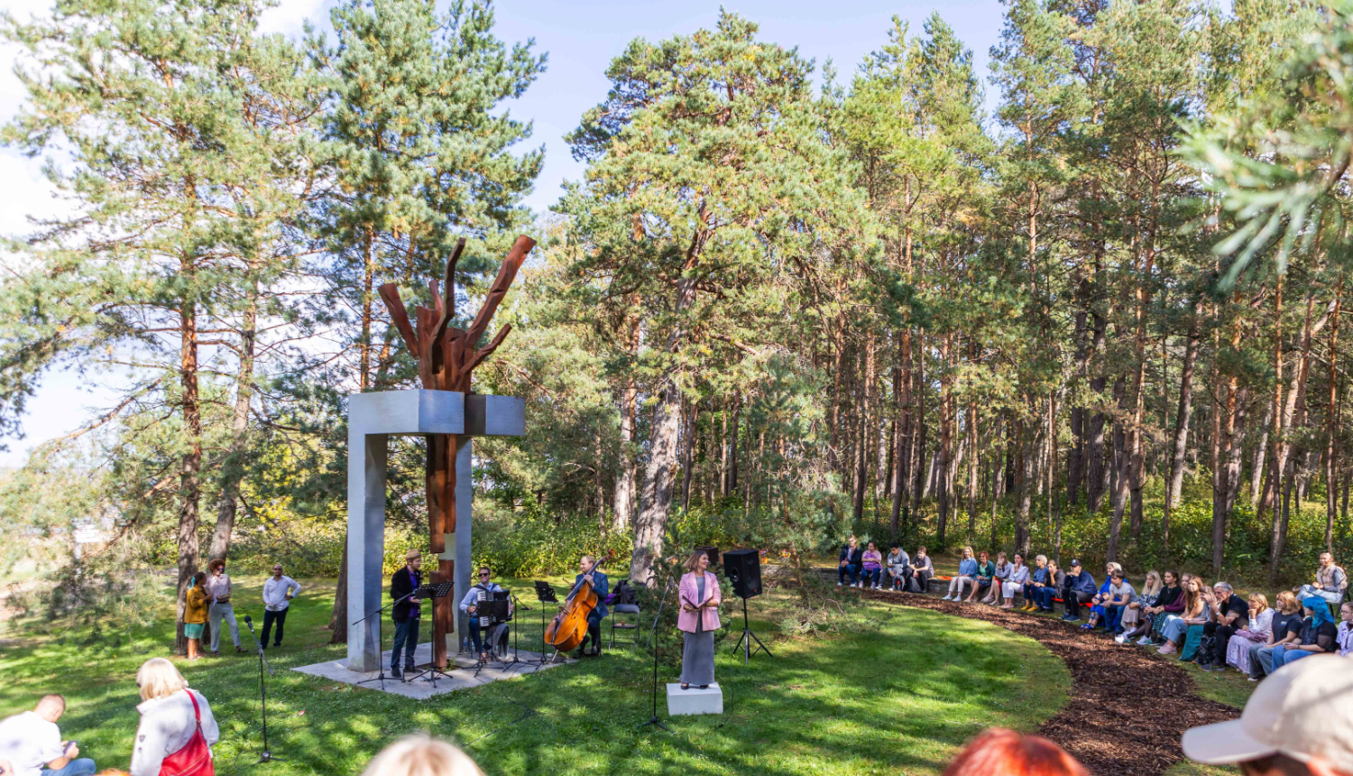 Pasākums brīvā dabā zem priedēm, kur skatītāji sēž puslokā uz koka soliņiem, klausoties priekšnesumā