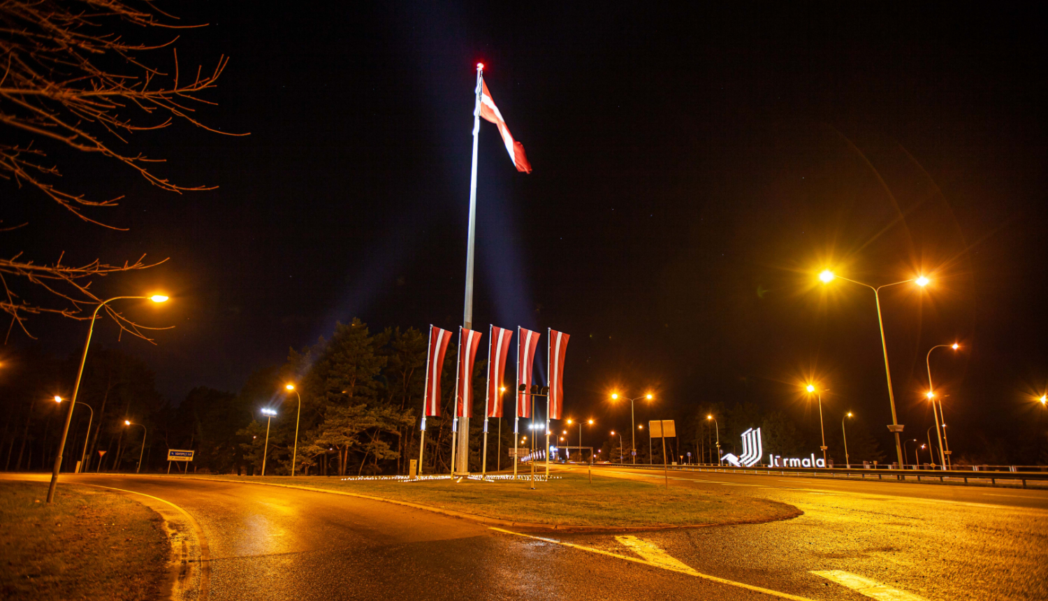 Nakts skats Jūrmalā: apgaismots satiksmes aplis ar paceltu Latvijas valsts karogu un vairākām mazākām karogmastiem svētku noformējumā pie Jūrmalas pilsētas robežas zīmes