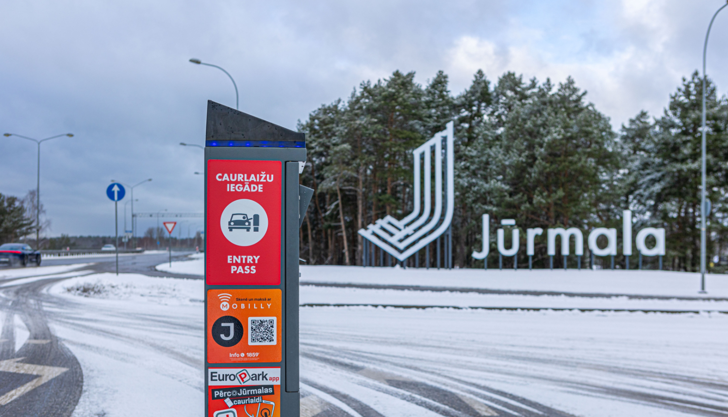 Ziemas ainava pie Jūrmalas pilsētas robežas. Priekšplānā redzama informatīva stenda zīme ar uzrakstu “Caurlažu iegāde / Entry Pass”, norādot, ka iebraukšanai Jūrmalā nepieciešama maksas caurlaide. Zemāk redzami dažādi maksāšanas veidi – Mobilly, EuroPark lietotnes un QR kods. Fonā redzams liels, balts Jūrmalas pilsētas nosaukums ar stilizētu logotipu priežu meža fonā. Ceļš un apkārtne ir pārklāti ar sniegu, debesīs – ziemīgi apmācies laiks.