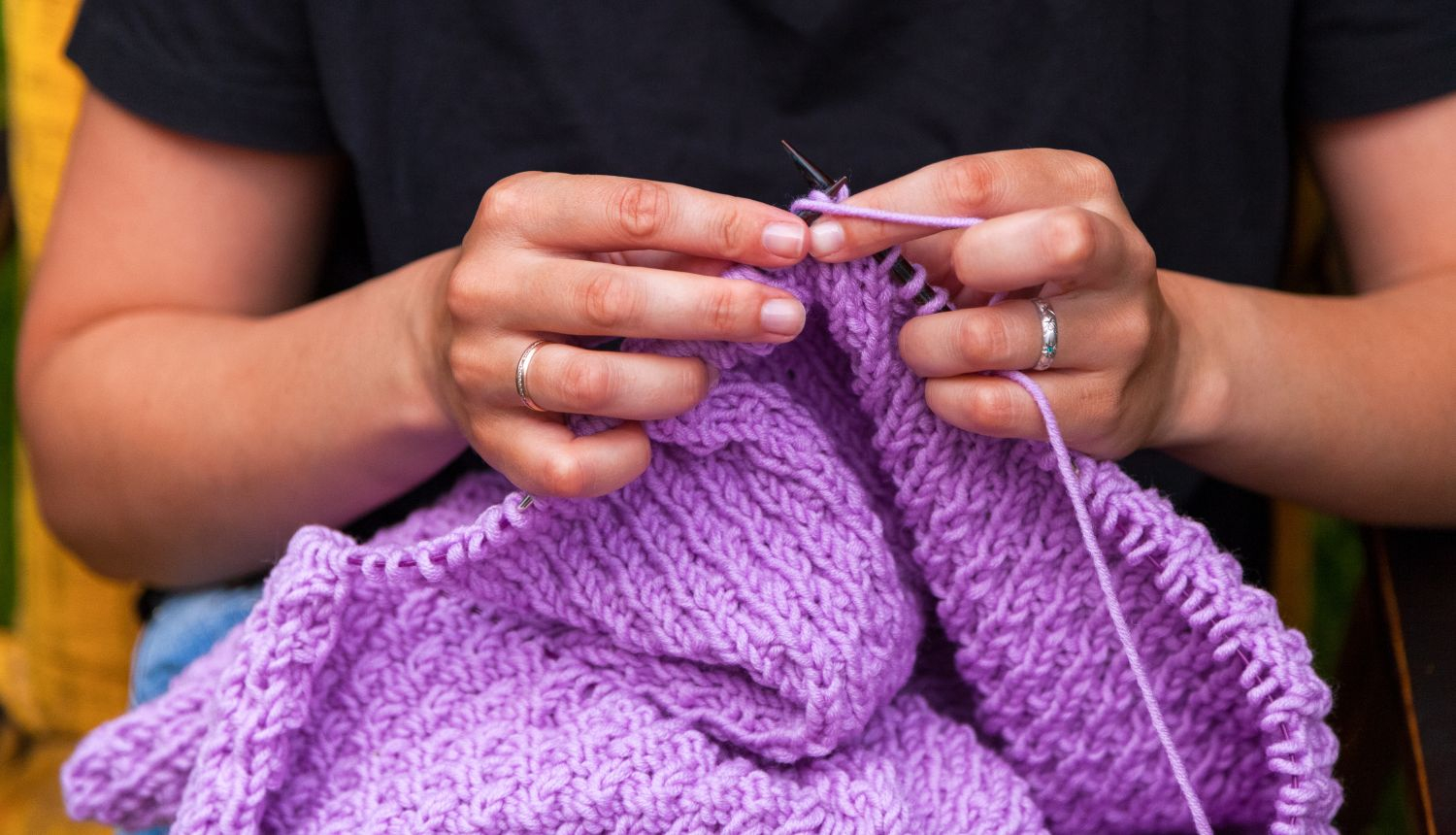 Persona ada ar adāmadatām no biezas, violetas dzijas, turot rokās veidojošos adījumu