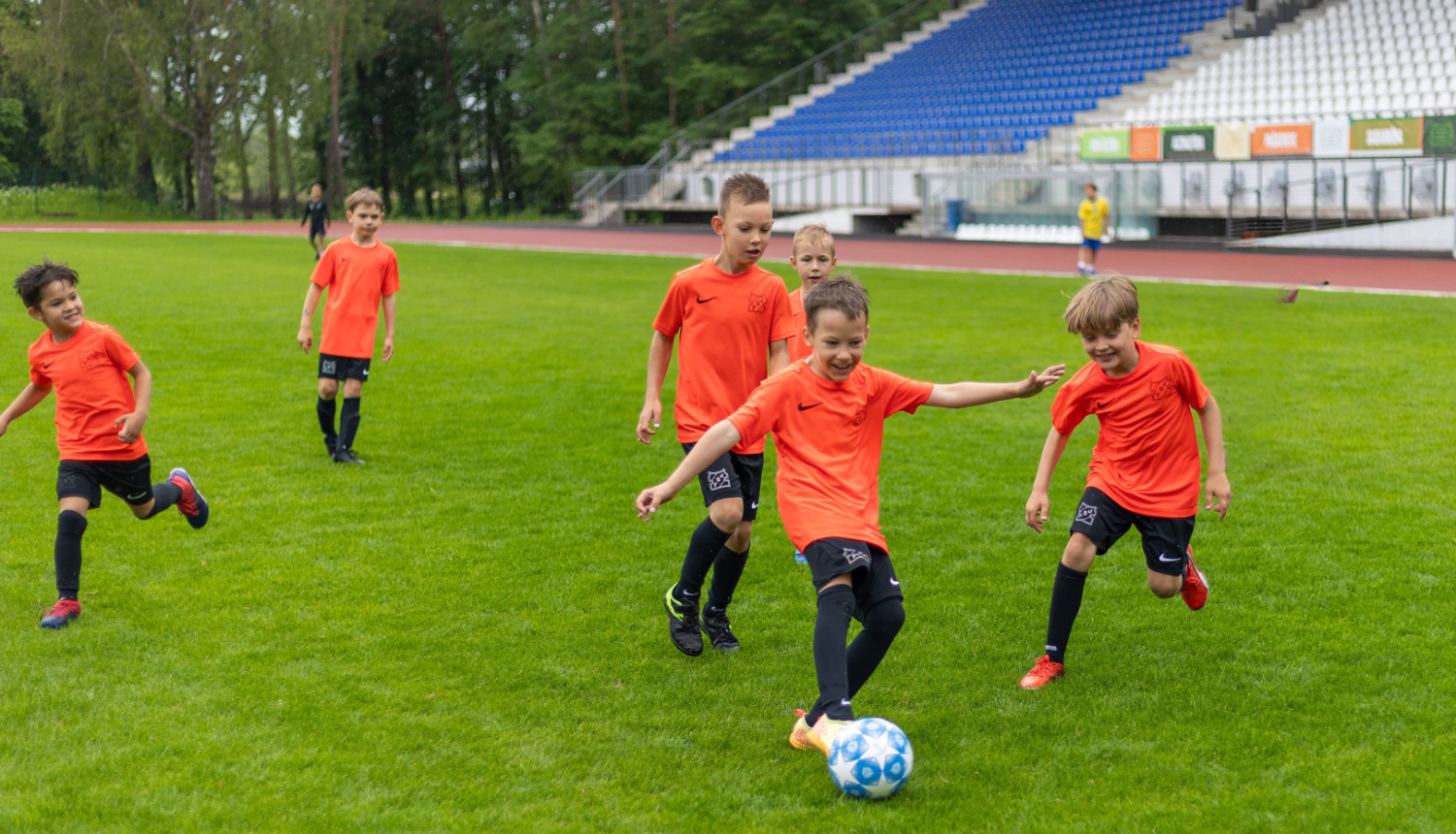 Bērni āra laukumā zaļā zālē spēlē futbolu