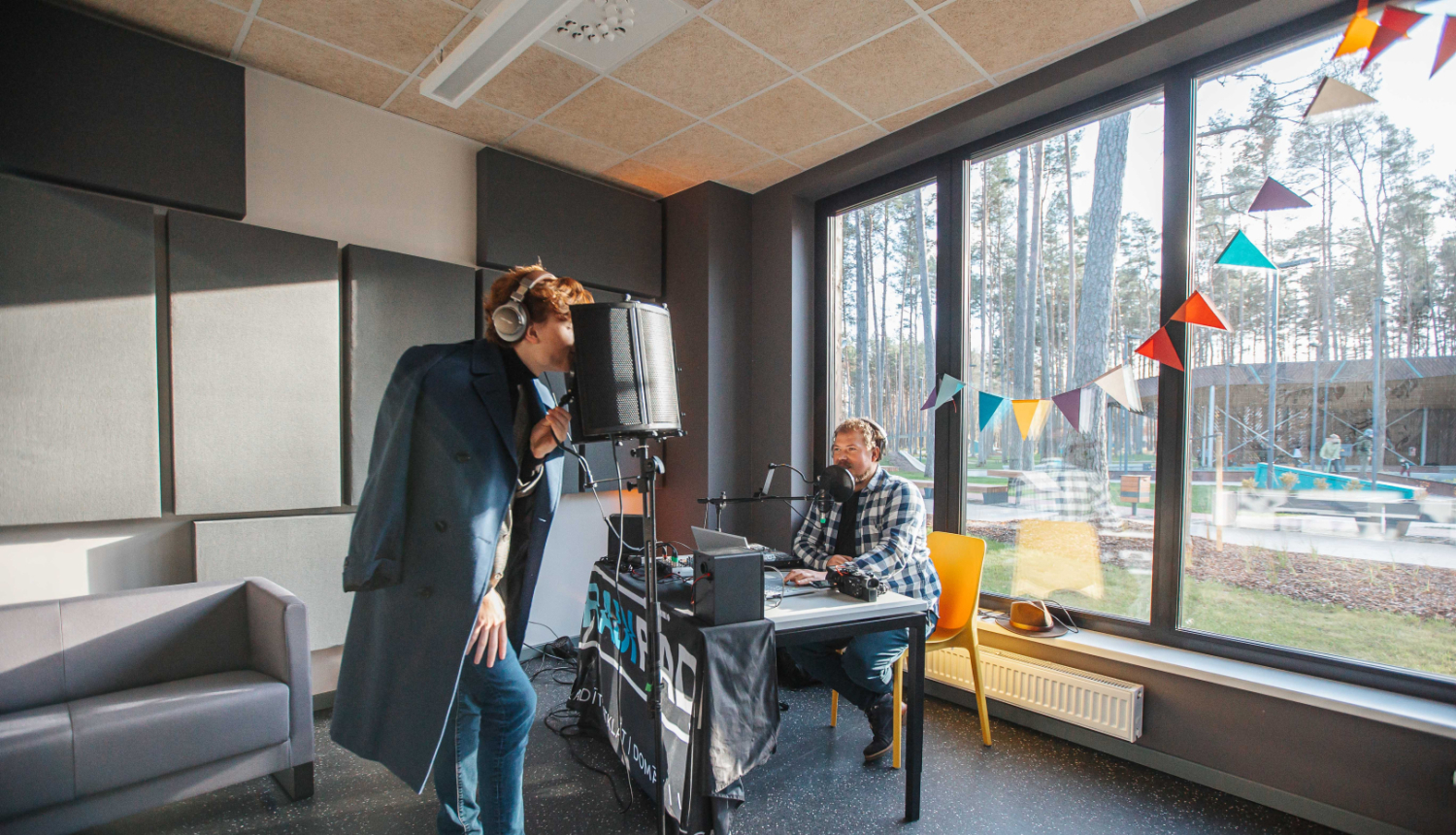 Divi jaunieši ieraksta audio materiālu modernā ierakstu studijā ar lieliem logiem. Viens stāv pie mikrofona ar austiņām un mēteli, otrs sēž pie galda ar datoru un audio aprīkojumu