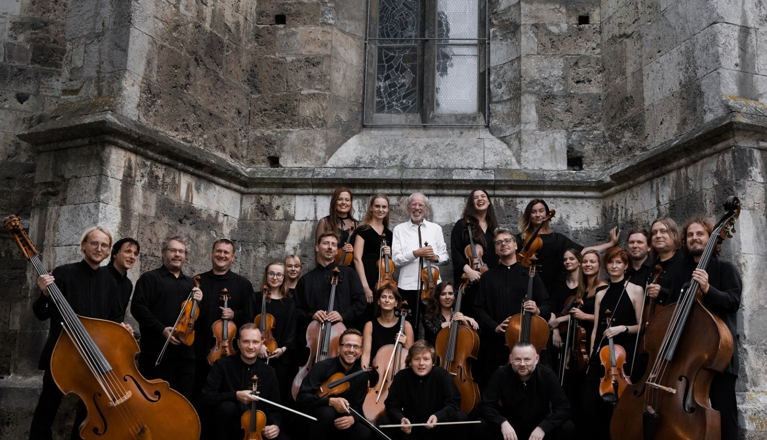 Grupas portrets ar klasiskā stila stīgu orķestri – apmēram 25 mūziķi pozē ārpusē pie vēsturiskas akmens ēkas sienas