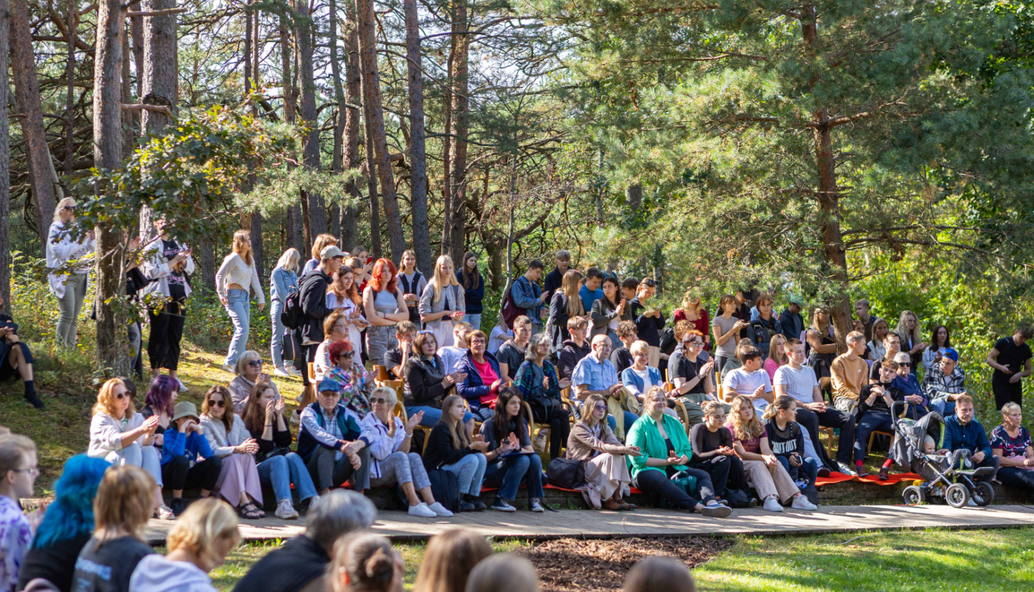 cilvēku grupa dažāda vecuma pārstāvniecībā sēž un stāv meža ielokā brīvdabas pasākumā, baudot Dzejas dienu norisi saulainā dienā
