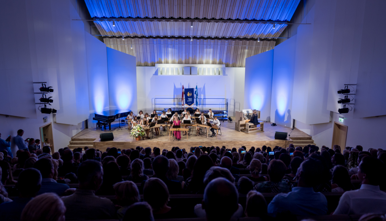 Koncertzāles skats no skatītāju rindām: uz skatuves uzstājas bērnu vai jauniešu kokļu ansamblis, fonā Latvijas un Jūrmalas karogi, labajā pusē redzams ērģelnieks