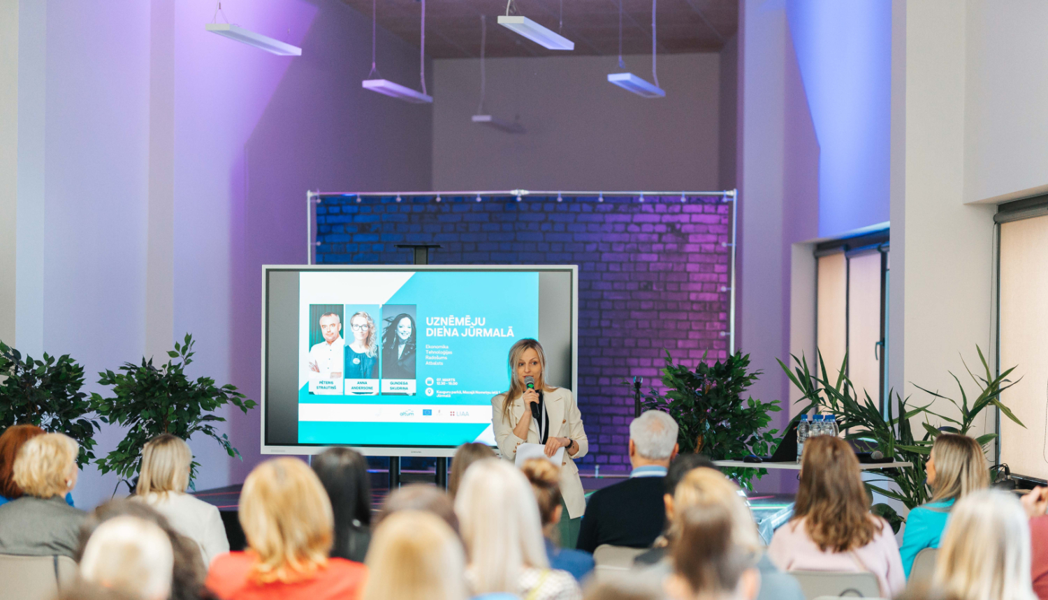 Konference ar nosaukumu 'Uzņēmēju diena Jūrmalā', kurā sieviete ar mikrofonu runā auditorijas priekšā