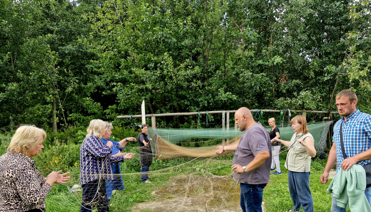 Cilvēku grupa brīvā dabā piedalās tradicionālā zvejas tīklu labošanas darbnīcā