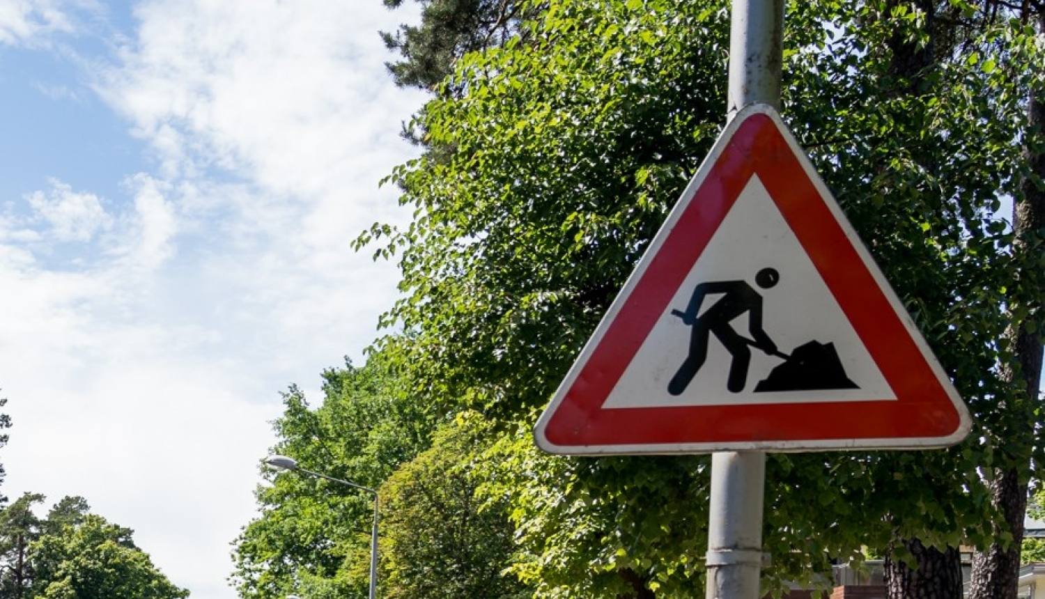 Ceļa zīme "Remonta darbi" pie ceļa, aiz zīmes redzami zaļi koki un zilas debesis ar dažiem mākoņiem